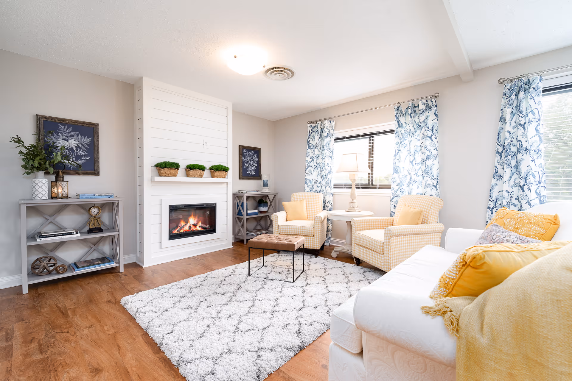 Bright and cozy living room with a white couch adorned with yellow pillows and a yellow throw blanket, two yellow checkered armchairs with yellow cushions, a white round side table with a lamp, a white and gray patterned rug on wooden flooring, a modern white fireplace with three small potted plants on the mantel, two gray shelves with decorative items, and windows with blue and white floral curtains letting in natural light.