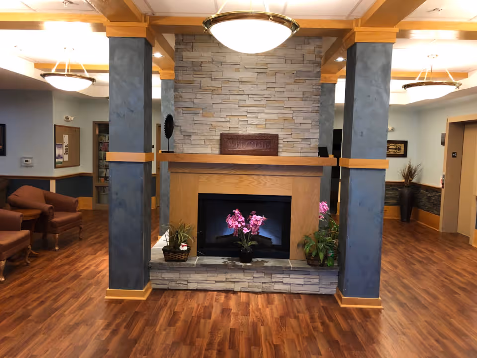 A senior living facility lobby with a central stone fireplace between pillars, wood floors, and seating.