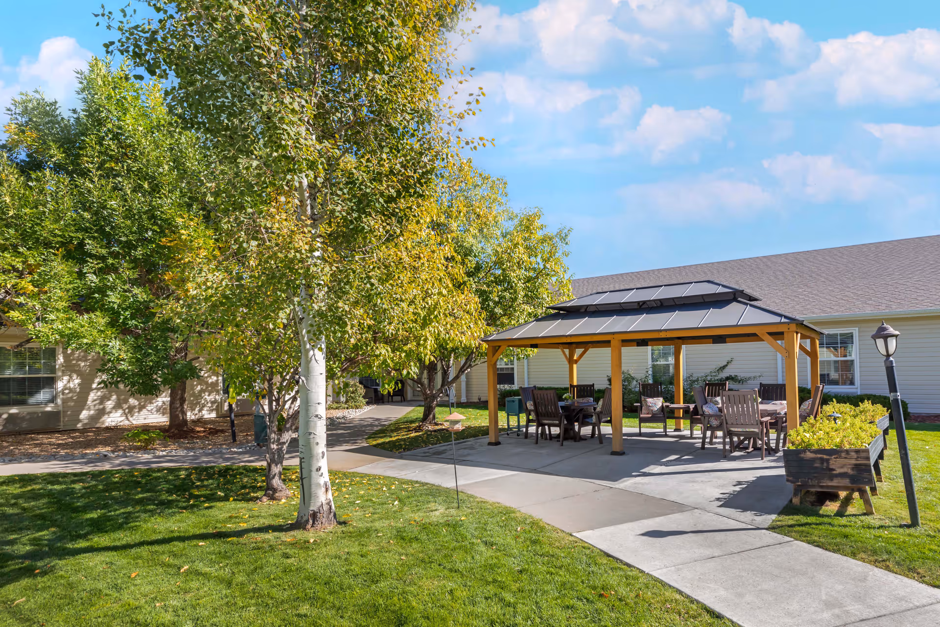 Outdoor seating area with a wooden gazebo covering several chairs and tables, surrounded by green grass, trees, and a paved walkway. A building with beige siding and windows is visible in the background under a partly cloudy blue sky.