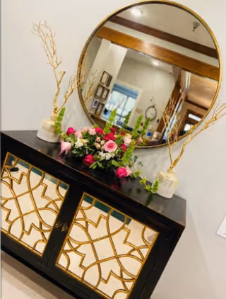A decorative black cabinet with intricate gold geometric patterns on the doors, topped with a floral arrangement featuring pink and white flowers and green foliage, flanked by two white vases holding tall, thin branches. Above the cabinet is a large round mirror reflecting part of the room, including a clock and framed pictures on the wall.