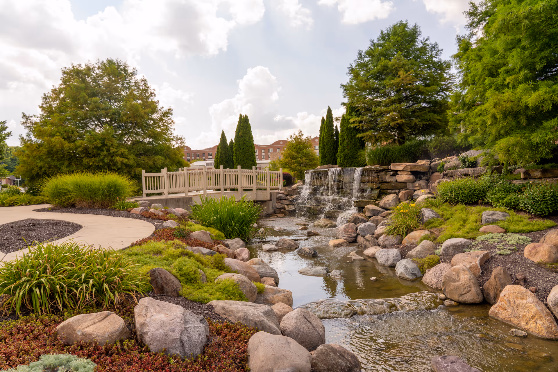 A landscaped outdoor area featuring a small waterfall flowing over rocks into a pond, surrounded by green shrubs, trees, and a walking path with a wooden bridge in the background under a partly cloudy sky.