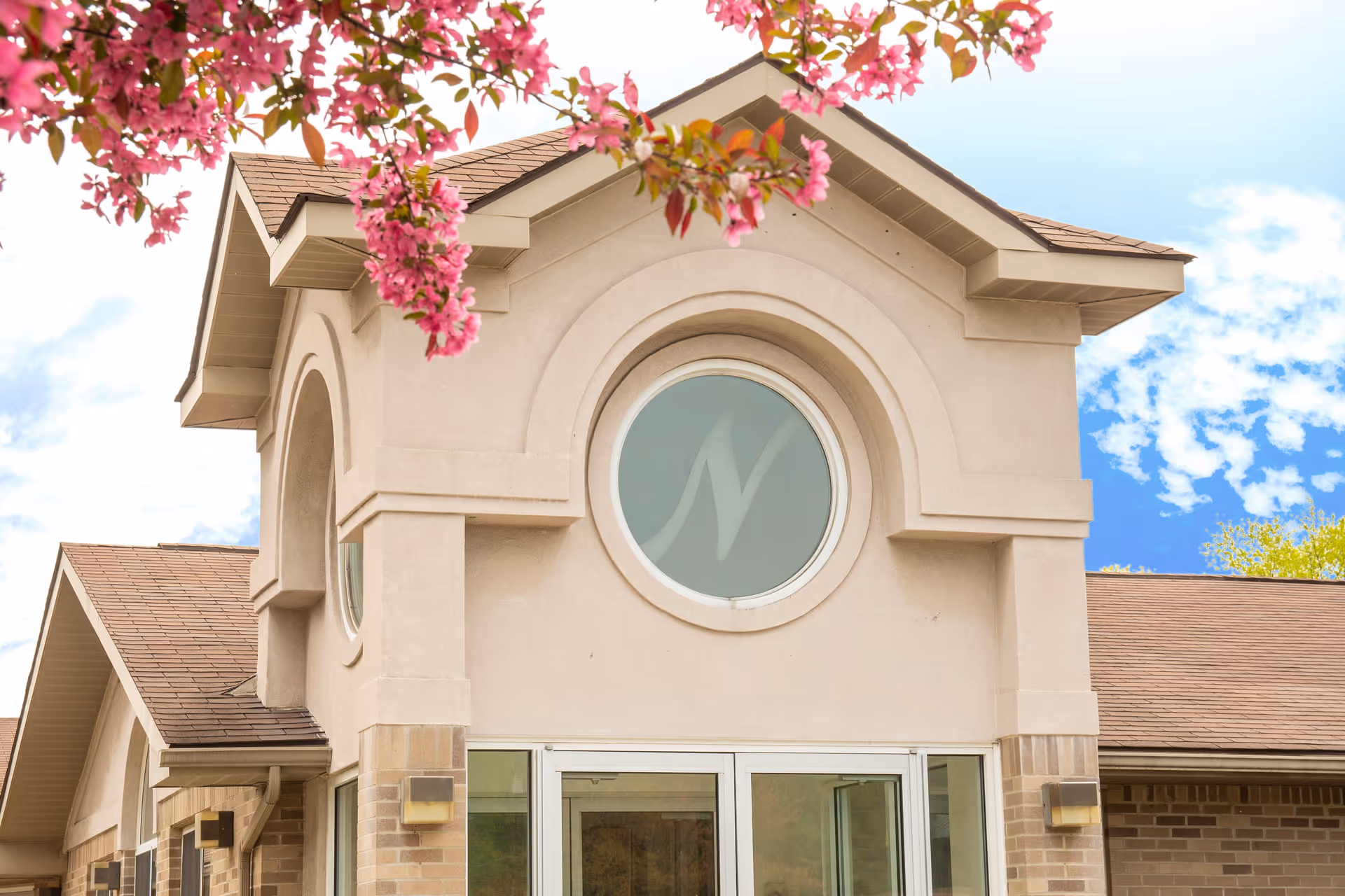 Exterior view of a building entrance with a round window above the door featuring a stylized 'N'. The building has beige walls, a brown shingled roof, and is framed by pink flowering tree branches in the foreground under a partly cloudy blue sky.