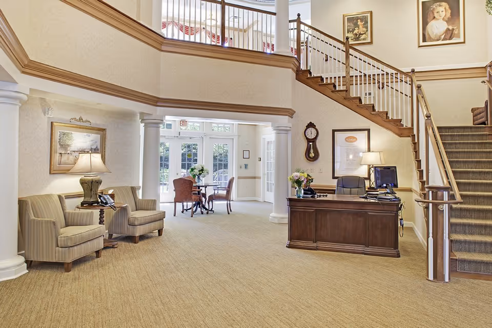 Reception lobby of a senior living facility with seating area, wooden front desk, staircase, and doors to an outdoor area.