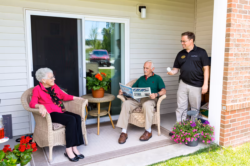 An elderly woman and an elderly man sitting on wicker chairs on a covered patio outside a building. The woman is wearing a bright pink top and black pants, smiling and looking at the man who is reading a newspaper. A younger man in a black shirt and beige pants stands nearby holding a white ball, smiling at the elderly man. There are potted flowers on a small table between the chairs and on the ground near the patio.