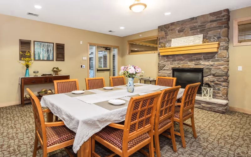 A dining room with a rectangular table covered with a white tablecloth and a runner, set with placemats and plates. Eight wooden chairs with patterned cushions surround the table. A vase with flowers is placed in the center. The room features a stone fireplace with a wooden mantel, a sideboard with decorative items, and a door leading outside. The walls are painted beige and the floor is carpeted.
