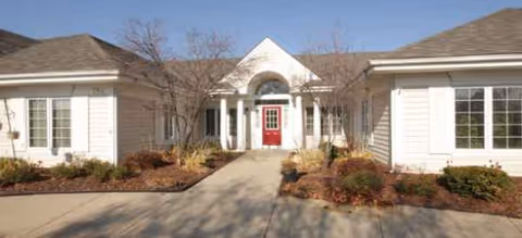 Front entrance of a light colored senior living building with a red door and arched portico.