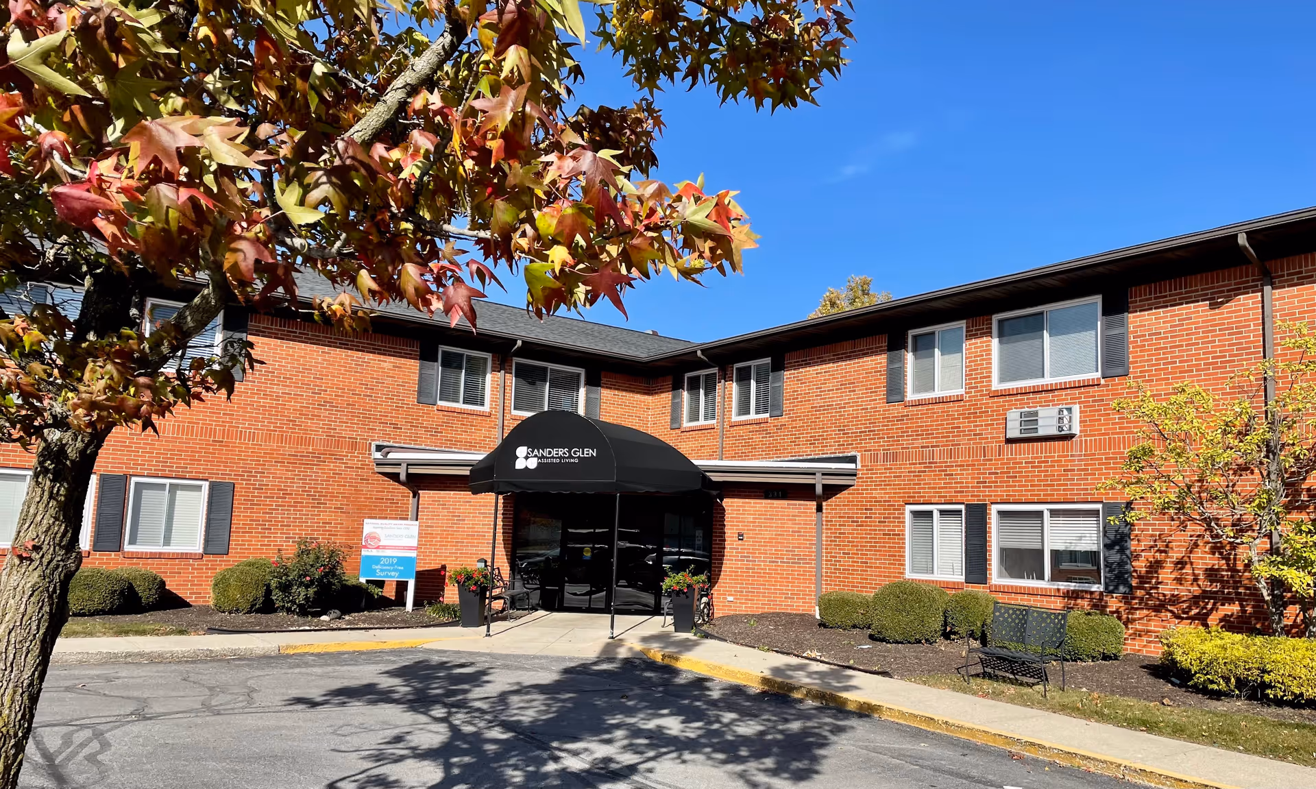 Exterior view of Sanders Glen Assisted Living Center, a two-story red brick building with black shutters and a black awning over the main entrance. There is a tree with autumn-colored leaves in the foreground and some bushes and a bench near the building. The sky is clear and blue.