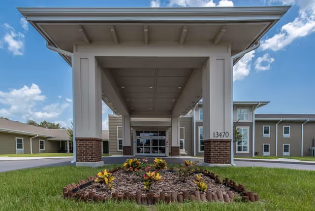 Front entrance of Twin Creeks Assisted Living & Memory Care facility with a covered driveway supported by large white columns with brick bases. There is a small landscaped area with plants and mulch in the foreground, and the building is a two-story structure with beige siding and multiple windows under a partly cloudy blue sky.