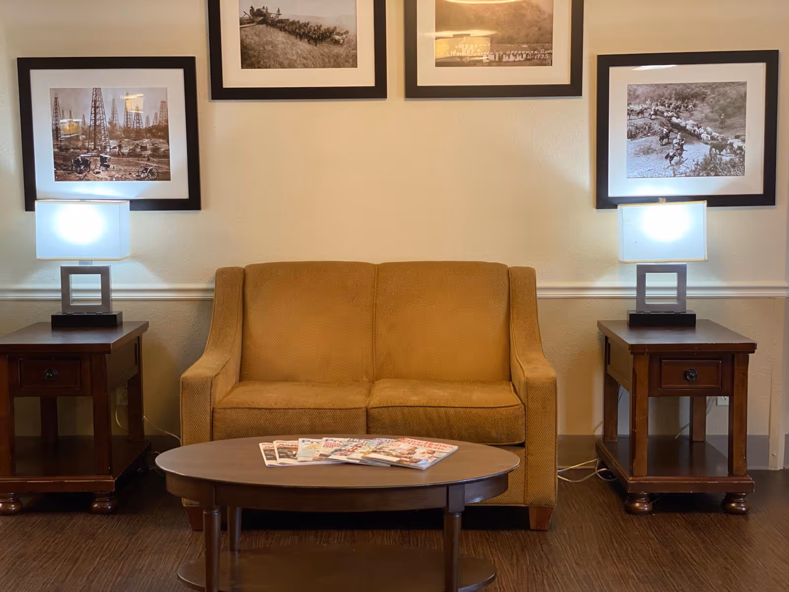 A cozy seating area with a mustard-yellow loveseat between two side tables topped with lamps, a coffee table with magazines in front, and framed photos on the wall.