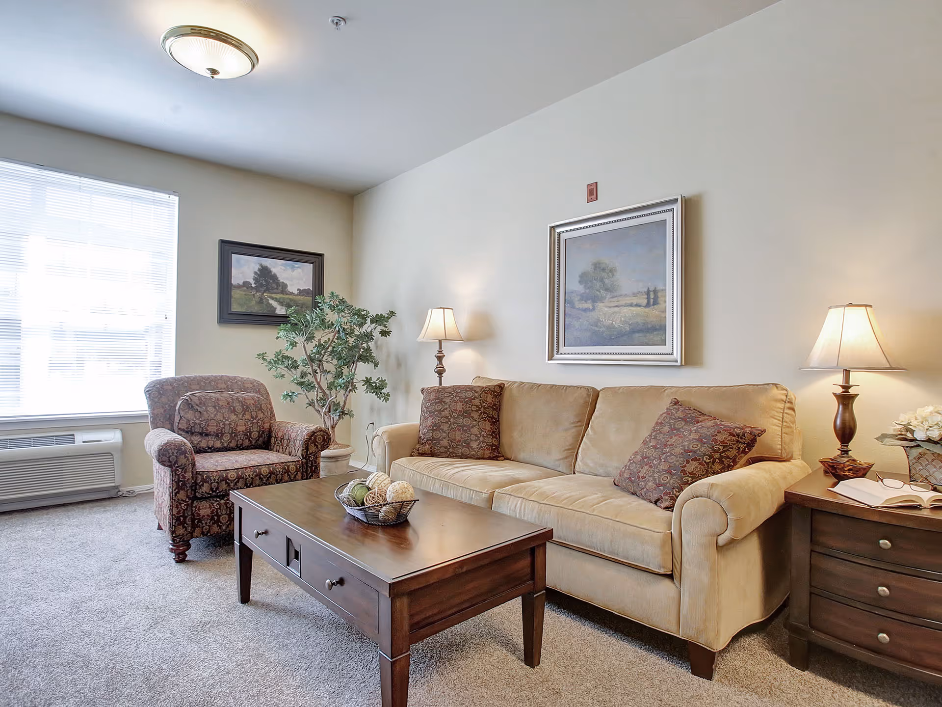 A cozy living room with a beige sofa adorned with patterned cushions, a matching armchair, a wooden coffee table with decorative balls in a basket, two table lamps on either side, a potted plant near the window, and framed landscape paintings on the walls.