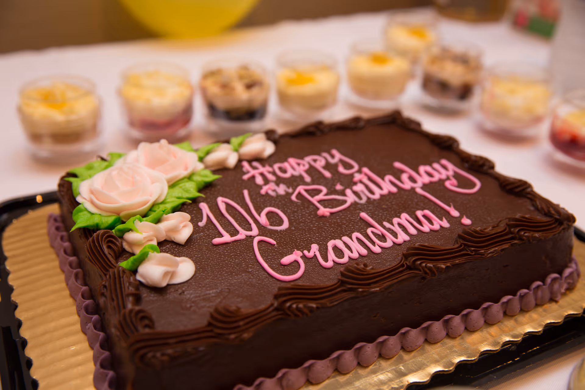 A decorated chocolate sheet cake with pink icing reading "Happy 106th Birthday Grandma!" and frosting roses on a table with small dessert cups behind it.