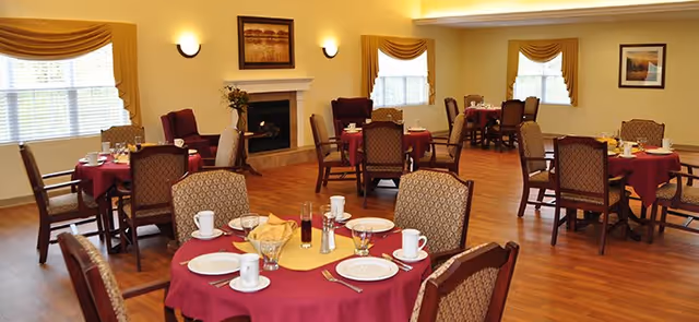 A dining room with multiple round tables covered with red tablecloths and set with white plates, cups, glasses, and silverware. The room has wooden flooring, beige walls, two windows with yellow curtains, a fireplace with a framed picture above it, and several upholstered chairs around the tables.