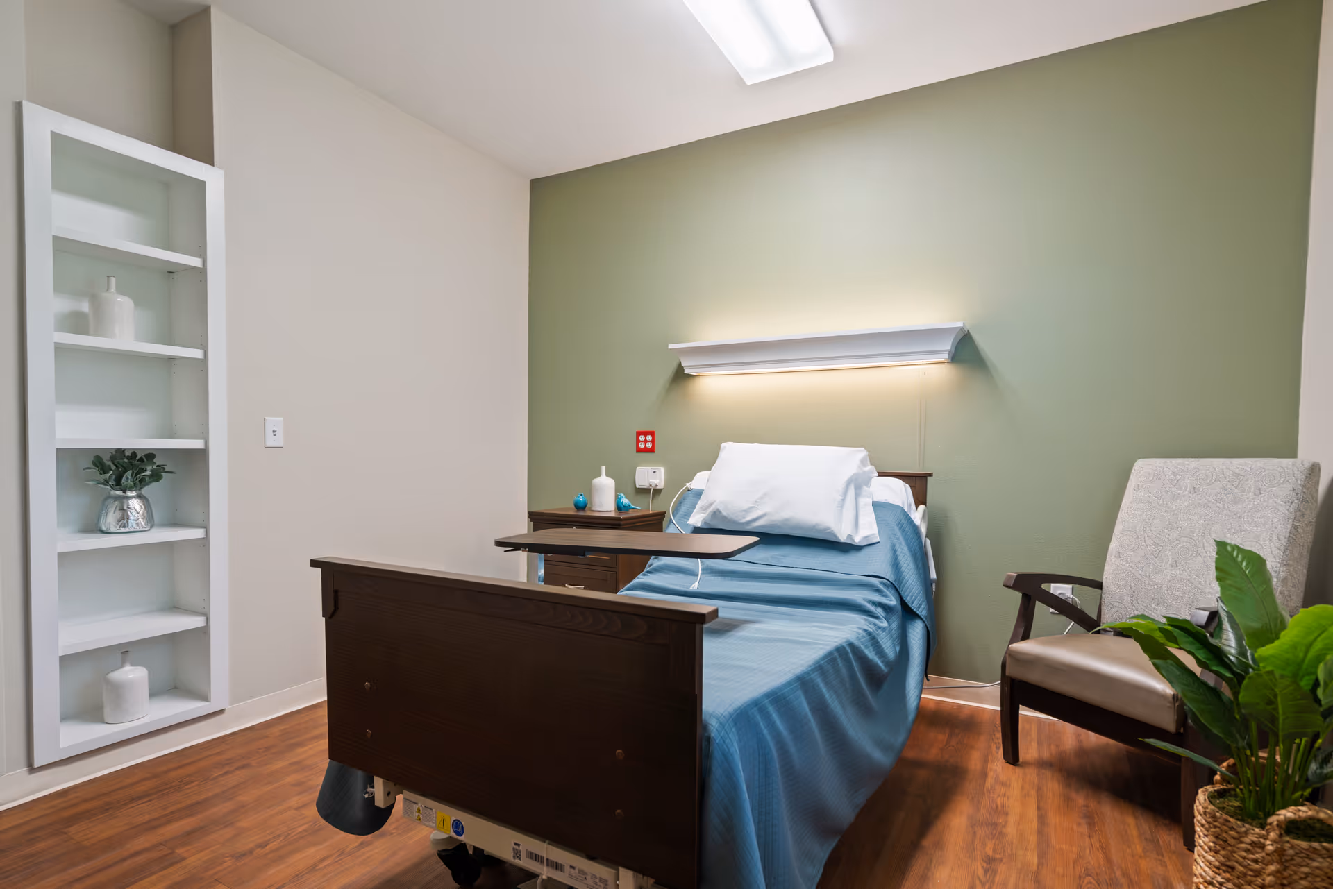 A hospital-style bed with blue bedding and a white pillow in a room with a green accent wall. There is a wooden nightstand with decorative items, a wall-mounted light above the bed, a cushioned chair, a potted plant, and a built-in white shelving unit with decorative vases.