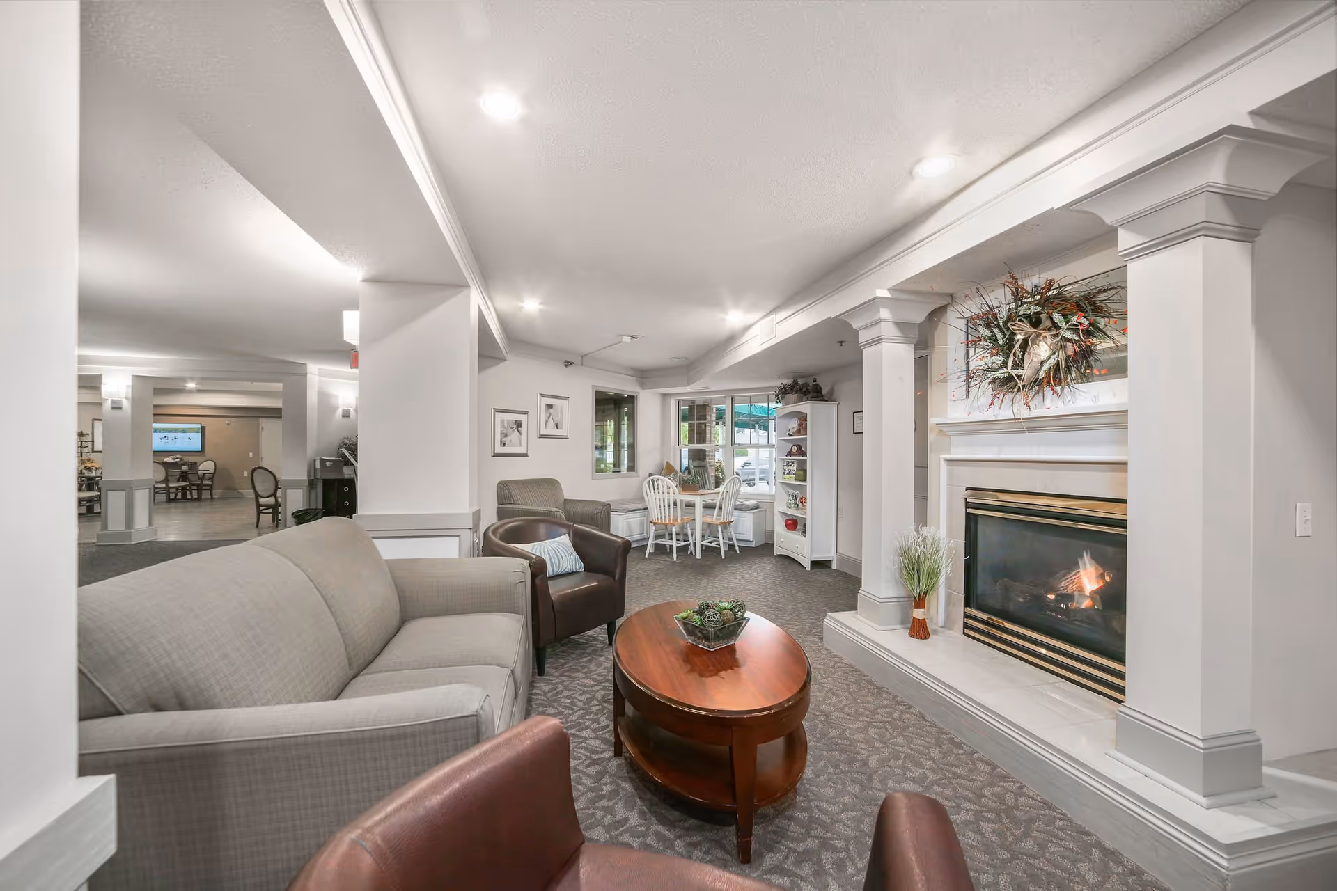A cozy senior living facility common area with a beige sofa, two brown leather chairs, a wooden oval coffee table, and a fireplace with a decorative wreath above it. In the background, there is a small dining nook with a round table and white chairs, a white shelving unit, and framed pictures on the wall. The space is well-lit with recessed ceiling lights and has carpeted flooring.
