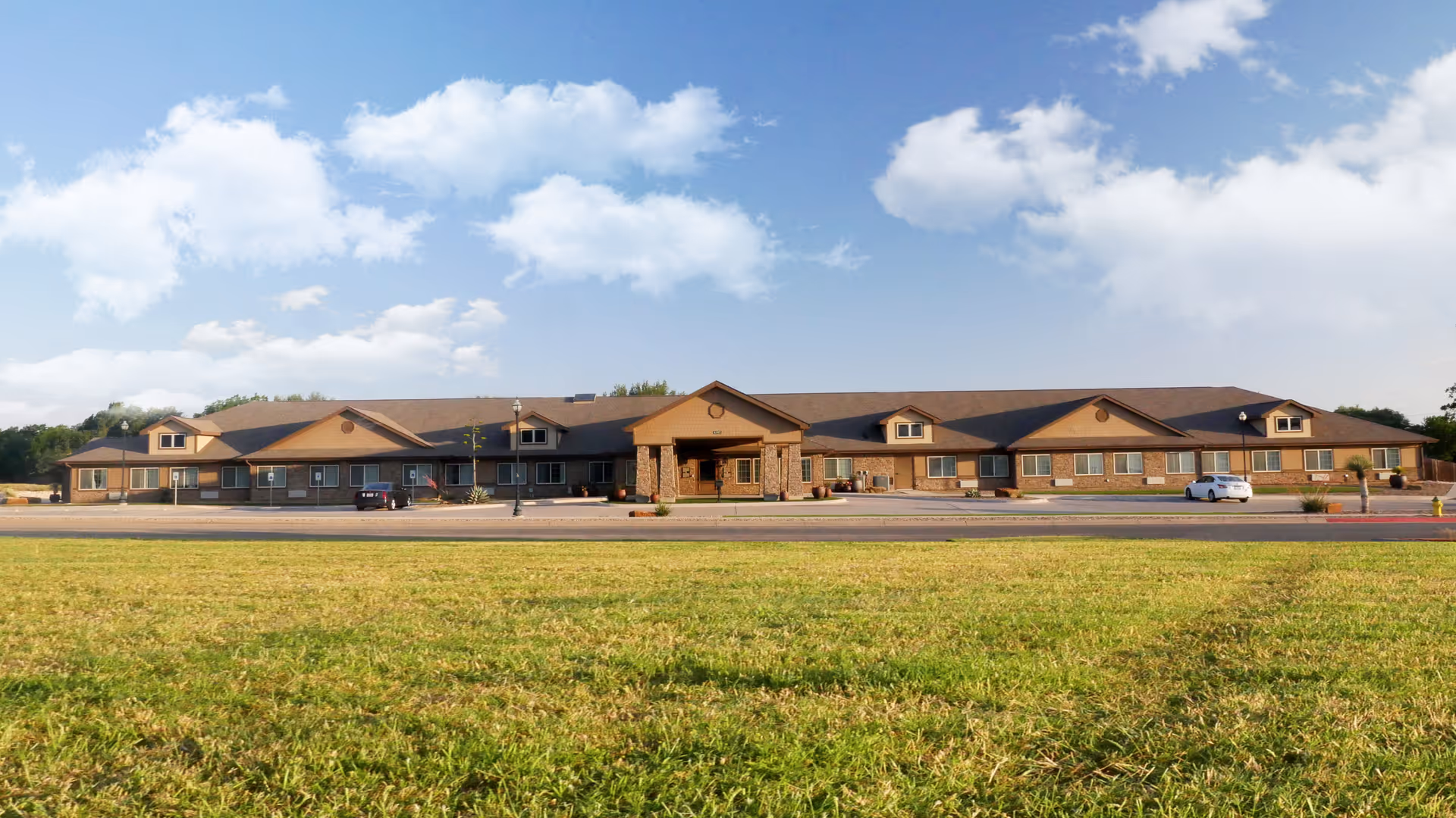 Wide exterior view of a single-story senior living facility building with a large grassy area in the foreground and a partly cloudy blue sky above. The building has a brown roof, stone pillars at the entrance, and several windows along the front. Two cars are parked near the building.