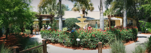 Outdoor garden area with a central three-tiered water fountain surrounded by lush green bushes with red flowers. Palm trees and other greenery are visible around the garden. There are pathways leading around the garden, and a few people are sitting on benches in the shaded areas near the building in the background.