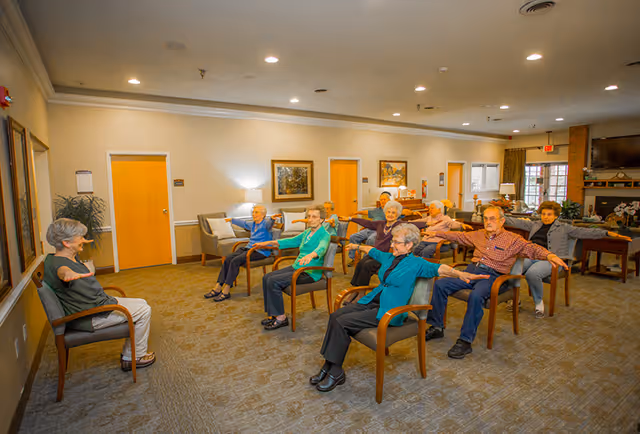 A group of elderly people seated in chairs arranged in a spacious room, participating in a seated exercise session led by an instructor also seated in a chair. The room has beige walls, several doors, framed artwork, and a television mounted on the wall.