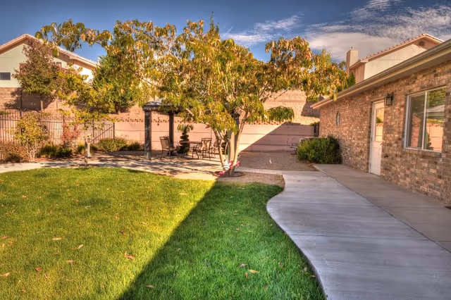 Outdoor area of a senior living facility with a green lawn, a tree with autumn leaves, a paved walkway, and a patio area with a pergola and outdoor seating. The building has brick walls and windows, and the sky is partly cloudy.