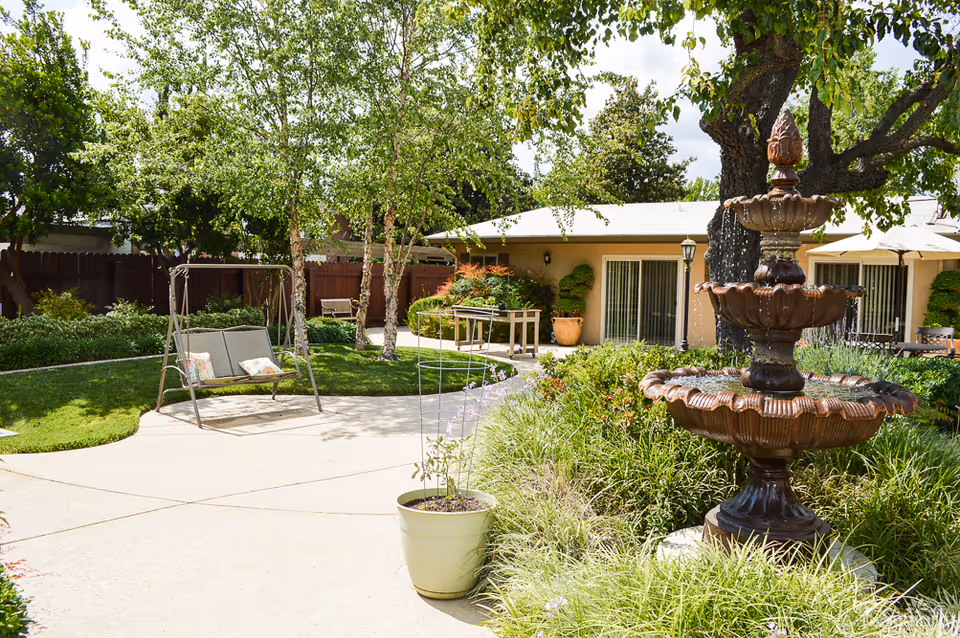 A sunny outdoor garden area at The WaterLeaf at Land Park featuring a large decorative three-tiered fountain surrounded by greenery and flowers. There is a paved walkway, a swing bench with cushions, several trees, and a building with sliding glass doors in the background.
