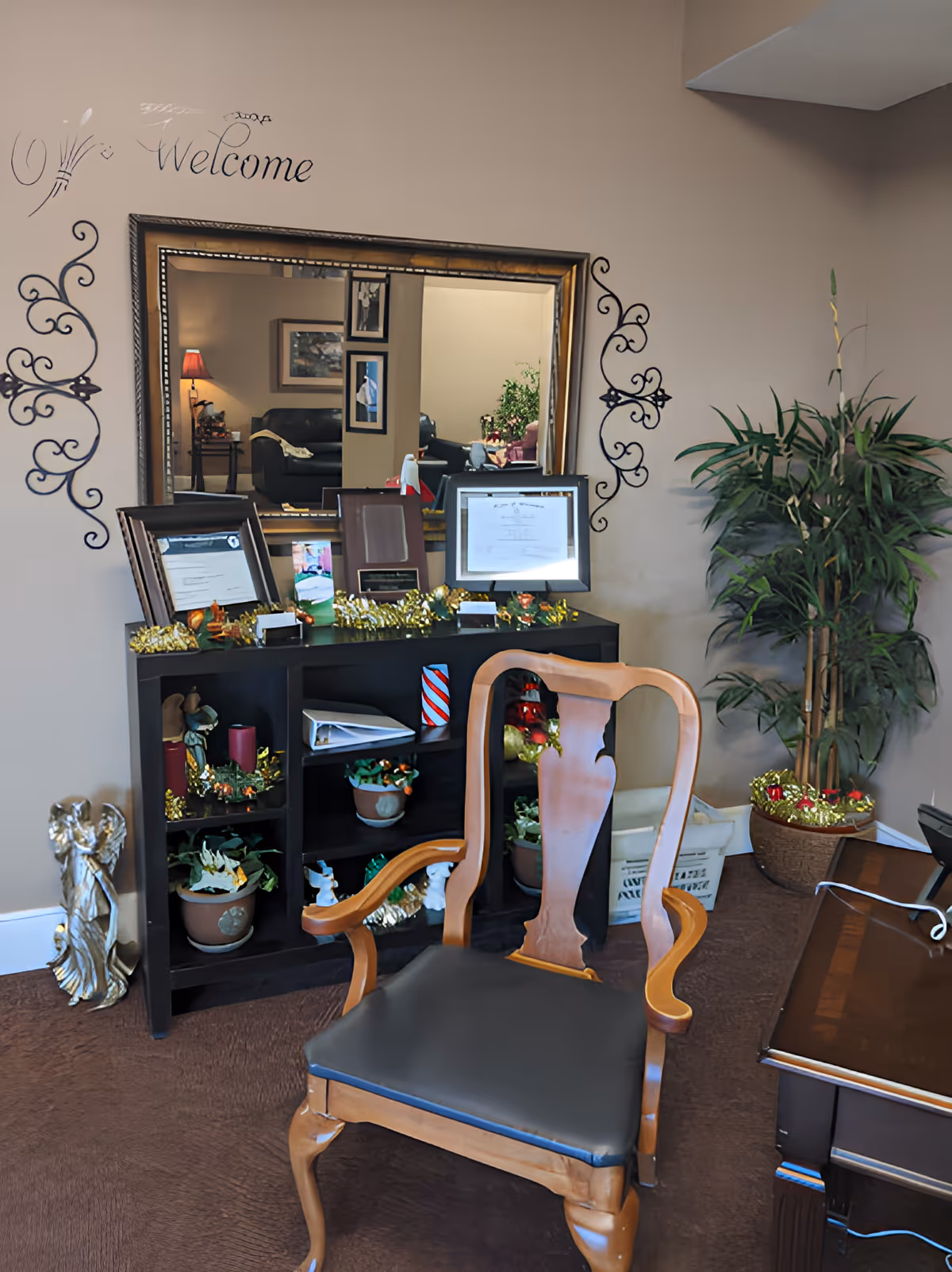A waiting-room style interior showing a wooden chair in front of a shelf and mirror decorated with framed certificates, plants, and holiday garlands.