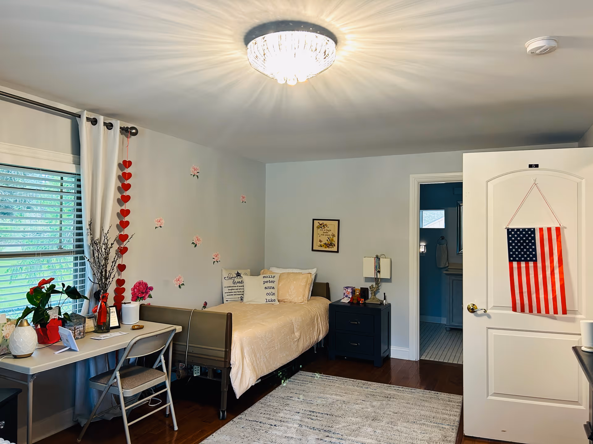 A cozy bedroom with a single bed covered in a beige quilt and decorative pillows. There is a black nightstand with a lamp and small items next to the bed. A white desk with a folding chair is placed near a window with white curtains and blinds. The wall above the bed is decorated with small flower decals and a framed picture. An American flag hangs on the inside of the open door, and a light fixture on the ceiling casts a soft glow over the room.