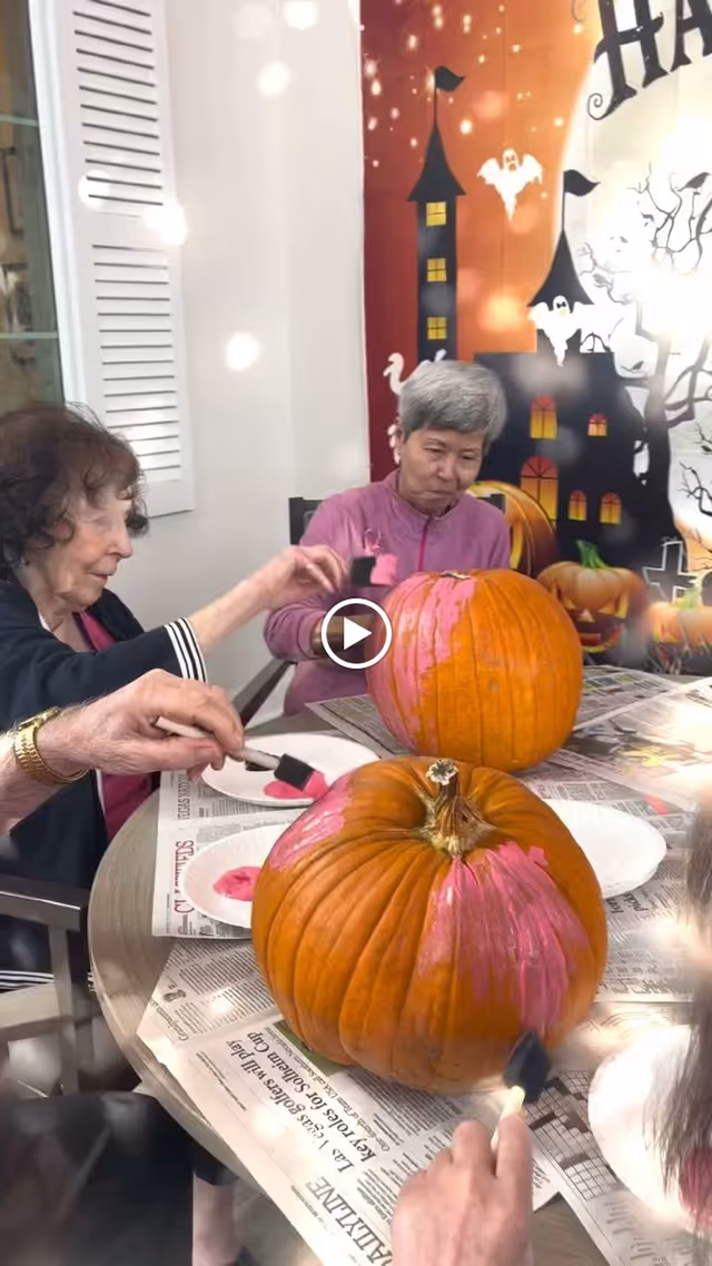 Three elderly women sitting around a table painting pumpkins with pink paint. The table is covered with newspapers and there is a Halloween-themed backdrop with haunted houses, ghosts, and pumpkins behind them.