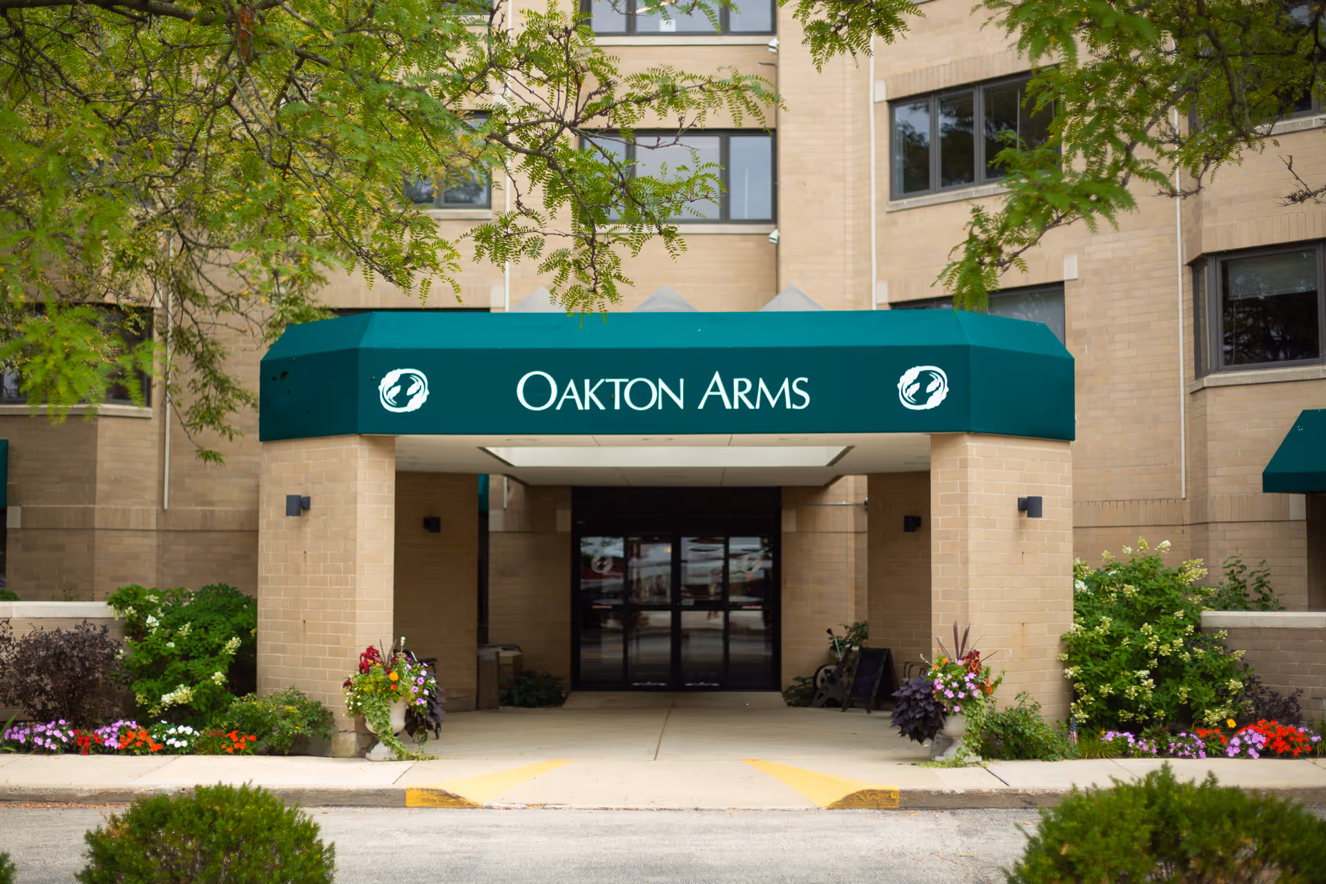 Entrance to Oakton Arms facility with a green awning displaying the name Oakton Arms, surrounded by beige brick walls and landscaped with colorful flowers and green bushes.