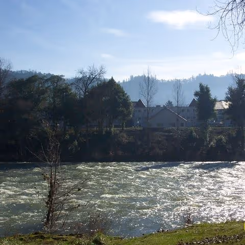 A flowing river with sunlight reflecting off the water, bordered by grassy banks and trees, with a building and forested hills in the background under a clear sky.