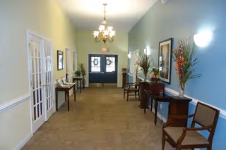 Entrance lobby hallway with double doors at the far end, chairs and decorative tables with framed art and floral arrangements along the walls.