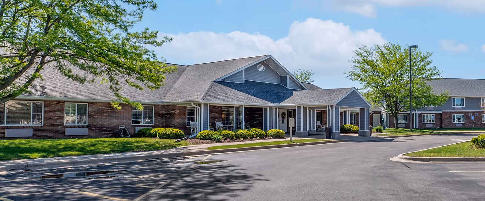 Exterior view of a senior living facility with brick and siding walls, a covered entrance, well-maintained landscaping including bushes and trees, and a paved driveway and parking area under a partly cloudy sky.