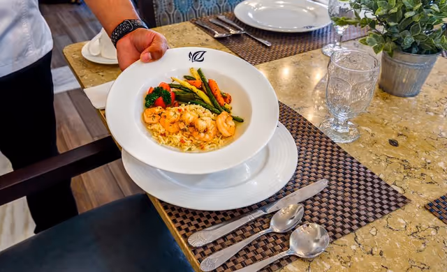 A person holding a white bowl filled with shrimp, rice, and mixed vegetables over a dining table set with plates, silverware, a glass, and a small potted plant.