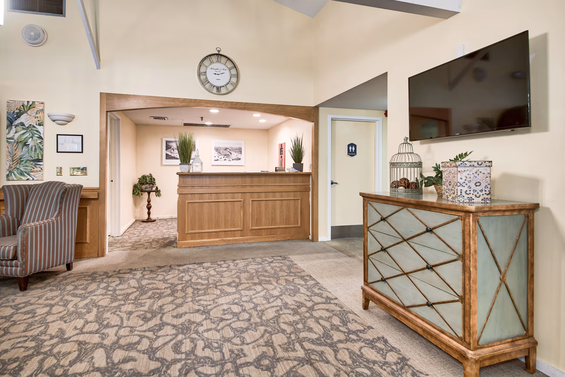 Interior view of a senior living facility reception area with a wooden front desk, a wall clock above it, and a door with a restroom sign to the right. There is a patterned carpet, a striped armchair on the left, and a wooden cabinet with decorative items and a mounted flat-screen TV on the right.