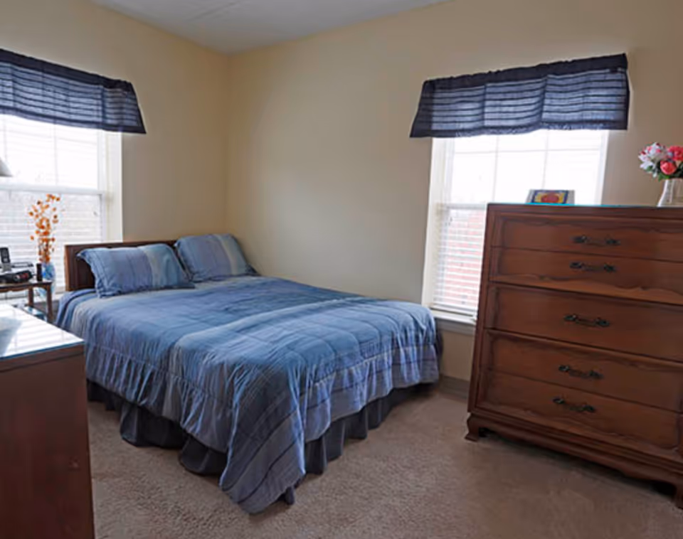 A bedroom with a neatly made bed covered in blue bedding, two windows with dark blue valances, a wooden dresser with drawers, and a small table with a vase of flowers and other items.