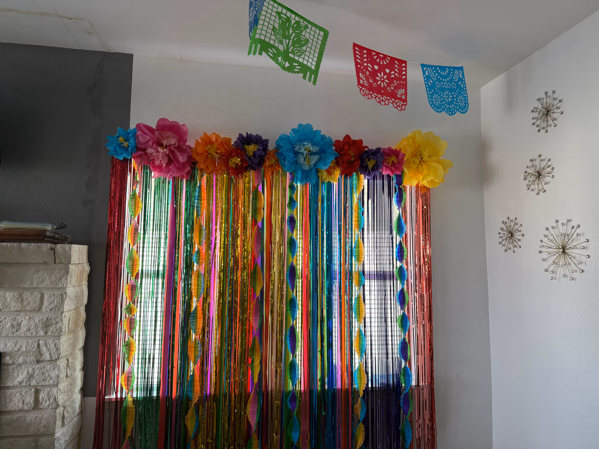 Interior living room window decorated with colorful metallic streamers, large paper flowers and papel picado banners next to a brick fireplace and wall art.
