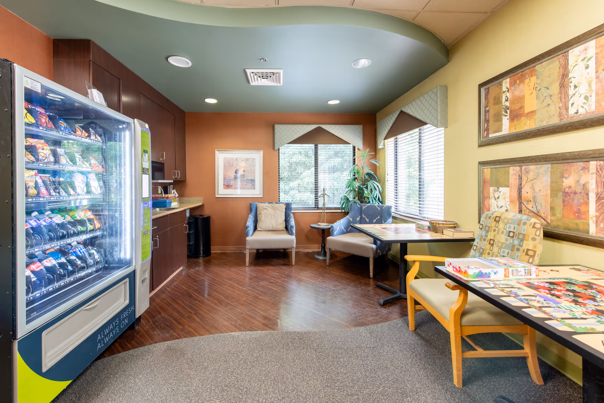 A cozy common area in a senior living facility with a vending machine stocked with snacks and drinks on the left. The room has wooden flooring and a green ceiling with recessed lighting. There are two armchairs and a small table near windows with blinds, and two tables with chairs on the right side, one with a puzzle and board game pieces on it. The walls are decorated with framed artwork and a potted plant is placed near the window.