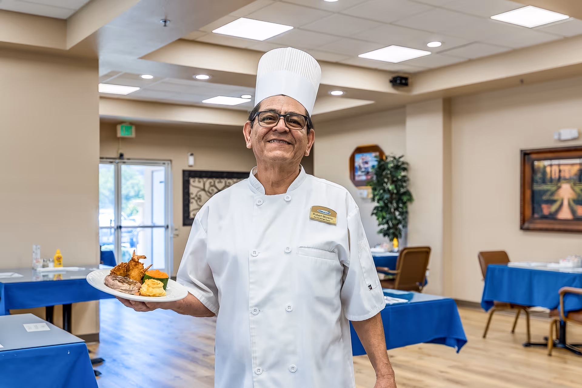 A smiling chef in a white uniform and tall hat holding a plated meal in a dining room with tables covered in blue tablecloths.