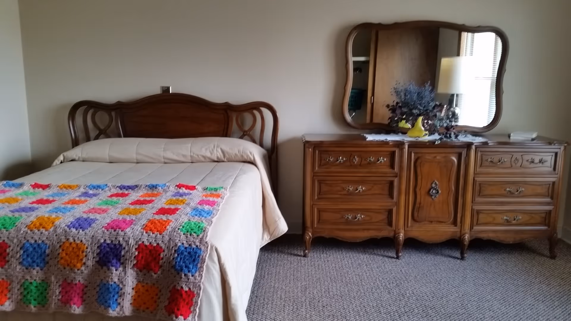 A bedroom with a wooden headboard bed covered with a beige comforter and a colorful crocheted blanket at the foot. Next to the bed is a wooden dresser with ornate handles and a large mirror above it. On the dresser is a vase with purple flowers and a table lamp. The room has beige walls and carpeted floor.