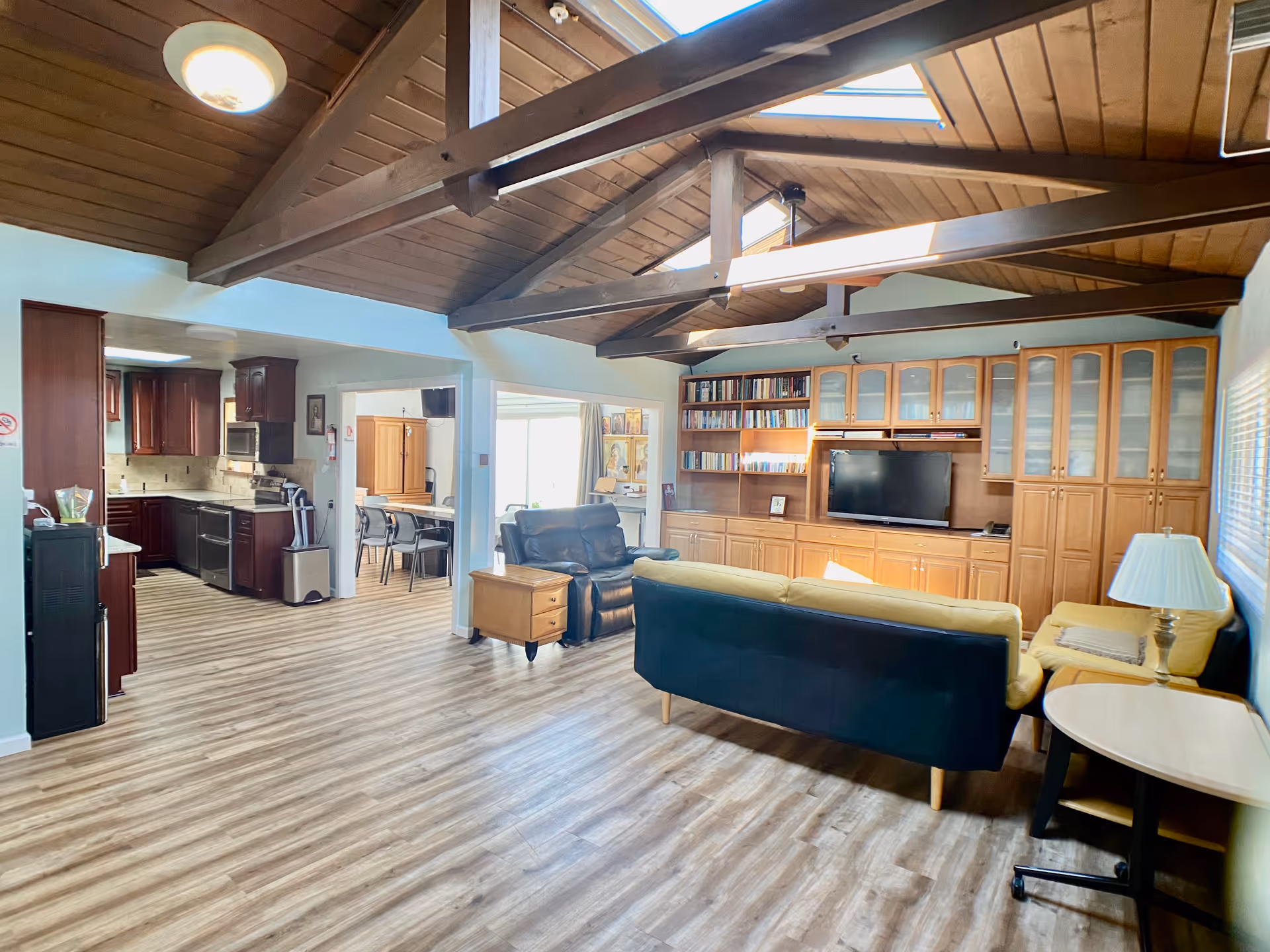 Interior view of a senior living facility common area with a wooden vaulted ceiling and exposed beams. The room features a living area with a black and yellow sofa, a black recliner, a wooden side table, and a lamp. There is a large wooden built-in cabinet with glass doors and shelves filled with books and a flat-screen TV. To the left, an open kitchen with dark wood cabinets and stainless steel appliances is visible, along with a dining area with a table and chairs. The floor is covered with light wood laminate.