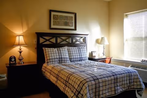 Sunlit bedroom with a plaid-covered bed, two bedside tables with lamps, framed art above the headboard, and a window with blinds.