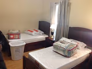 A simple bedroom with two single beds, each with a mattress and some bedding on top. There is a small nightstand with a lamp between the beds, a laundry basket on the floor, and curtains covering a window in the background.