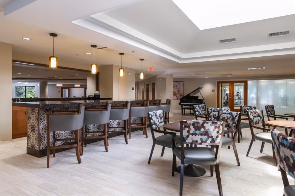 A spacious and well-lit common area with a bar counter featuring patterned tiles and several bar stools. There are multiple round tables surrounded by chairs with colorful hexagonal patterned upholstery. In the background, a black grand piano is visible near a set of glass doors, and the ceiling has recessed lighting and a tray design.