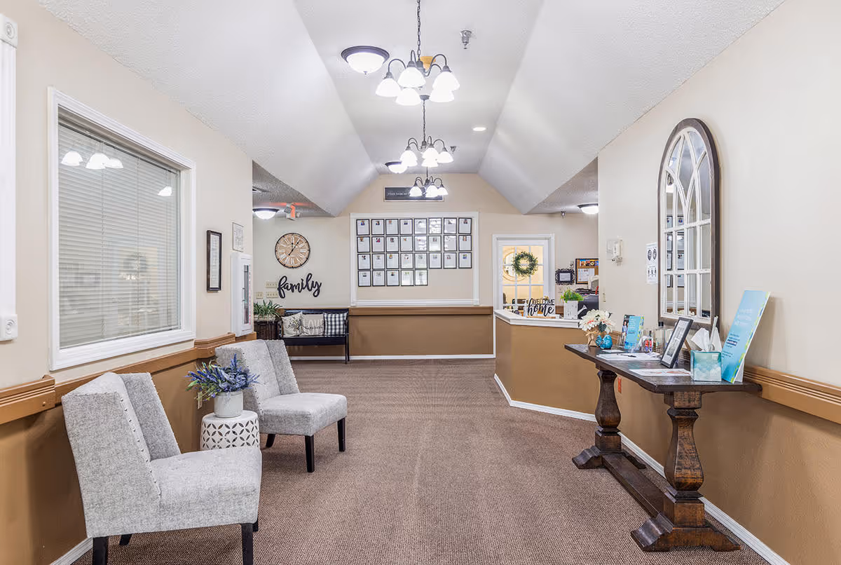 A well-lit interior hallway of a senior living facility with beige walls and brown carpet. Two gray upholstered chairs with a small white side table holding a vase with purple flowers are on the left. On the right, a wooden table displays informational brochures and decorative items beneath a large arched mirror. At the end of the hallway, a bench with pillows is placed under a wall clock and a sign that says 'family'. Multiple framed photos or certificates are displayed on the far wall above a reception counter with a wreath on the door behind it.