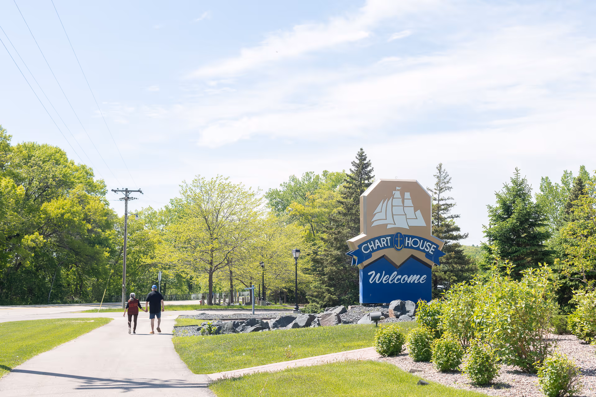 Two people walking on a paved pathway next to a large sign that reads 'Chart House Welcome' surrounded by green trees and bushes under a partly cloudy sky.