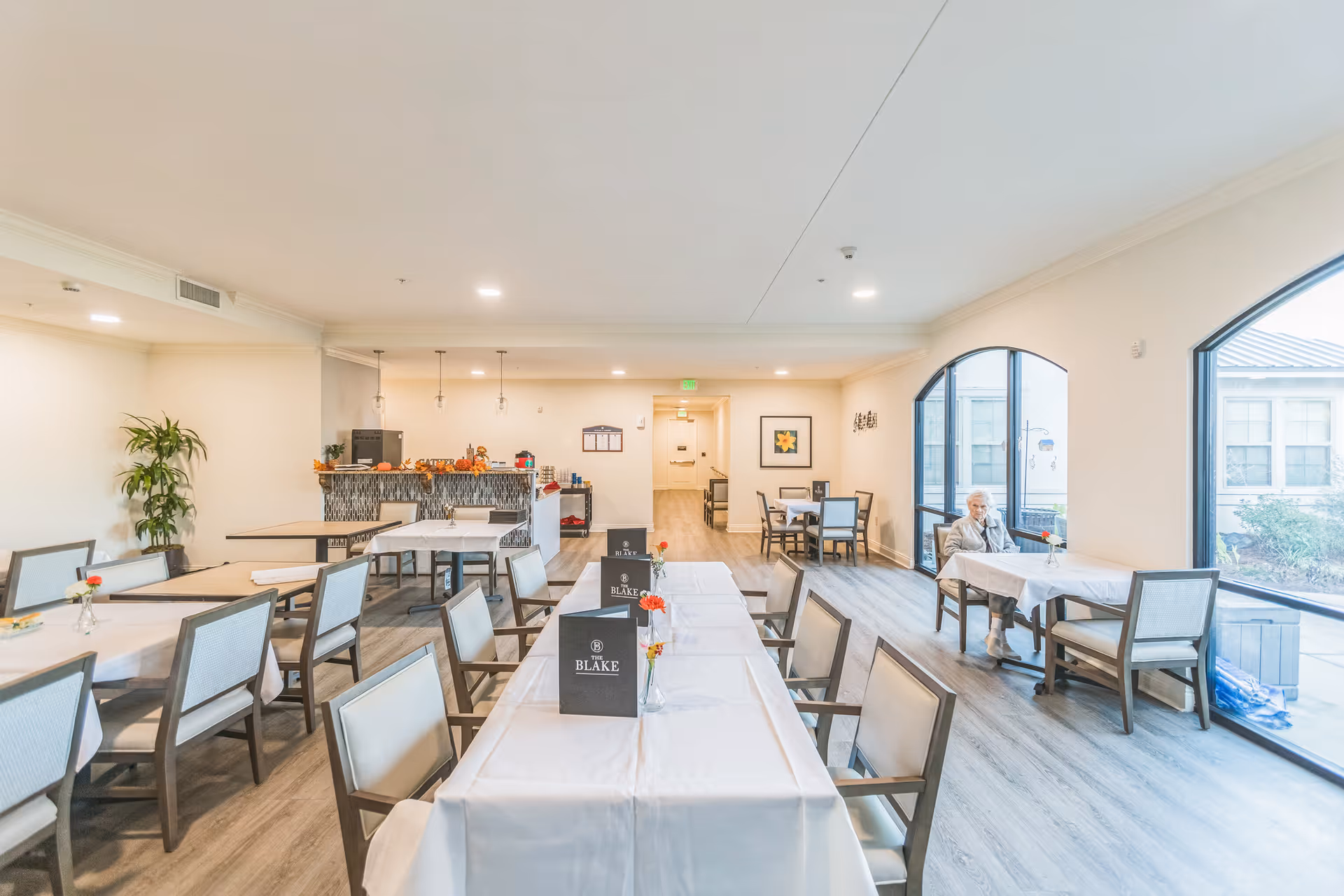 Bright dining room with long tables and chairs, a service counter in the back, and a person seated near large arched windows.