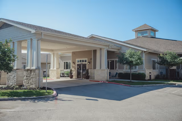 Exterior view of a single-story senior living facility building with a covered entrance supported by columns, surrounded by small trees and a paved driveway under a clear blue sky.
