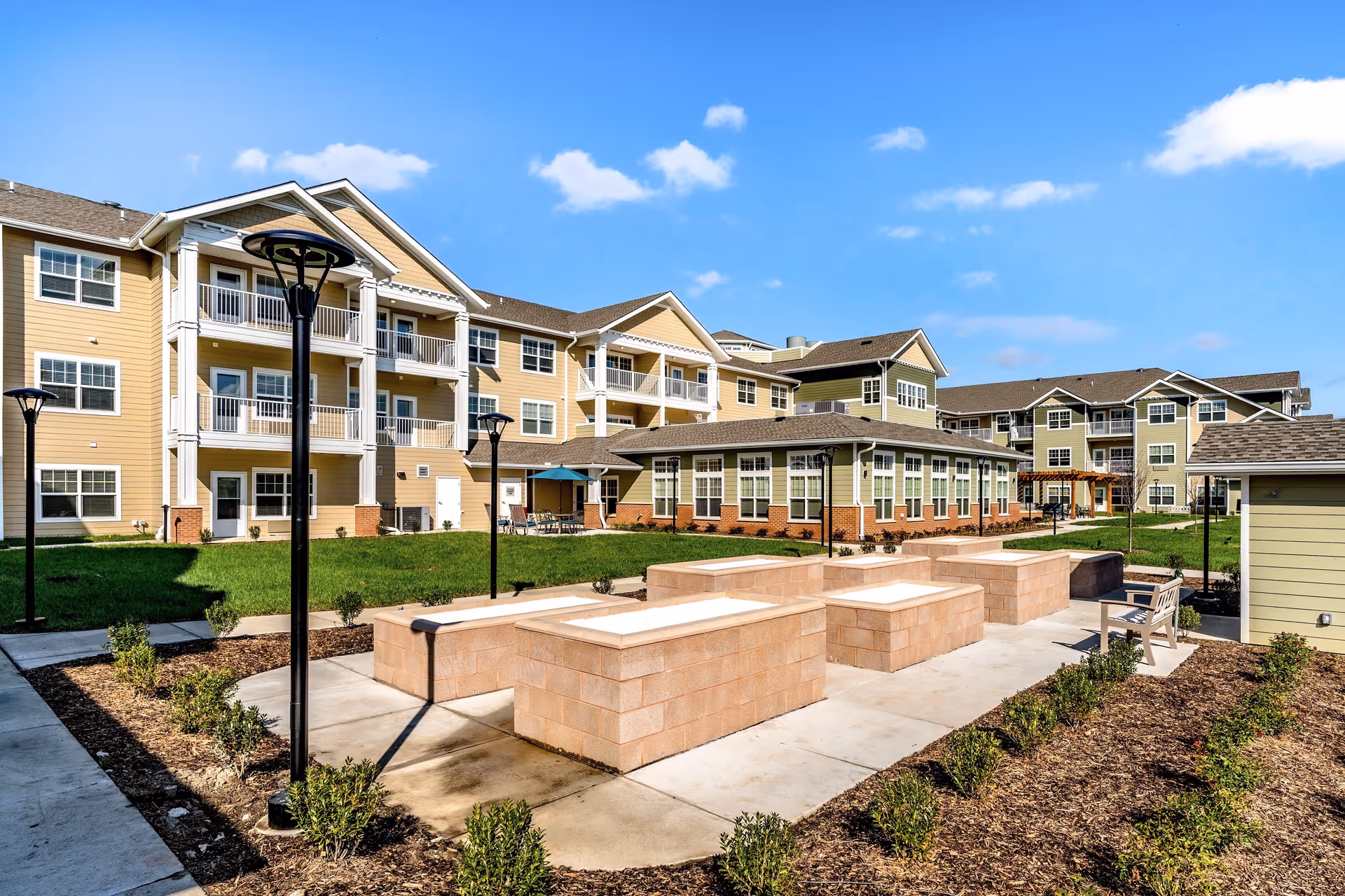 Outdoor courtyard area of Capitol Ridge Gracious Retirement Living featuring raised garden beds, benches, lamp posts, and a multi-story residential building with balconies in the background under a clear blue sky.