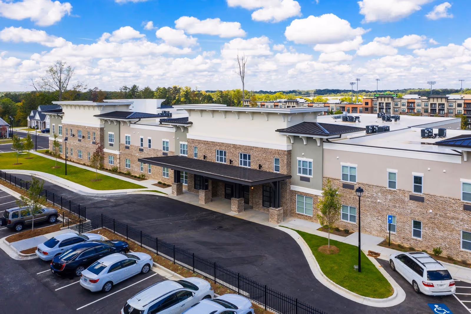 Front exterior of a three-story senior living facility with a covered entrance, parking lot, and landscaped grounds under a partly cloudy sky.
