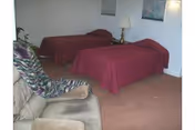 A room with two single beds covered with red bedspreads, a beige armchair with a patterned cushion, a small table with a lamp between the beds, and two framed pictures on the wall.