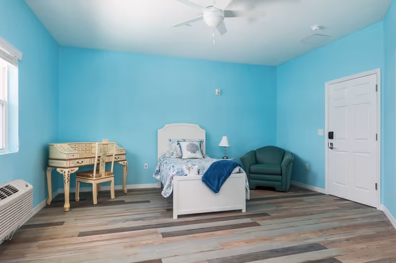 Bright turquoise-walled bedroom with a single white bed dressed in seashell bedding, a small decorative desk and chair, a green armchair, ceiling fan, window, and entry door.