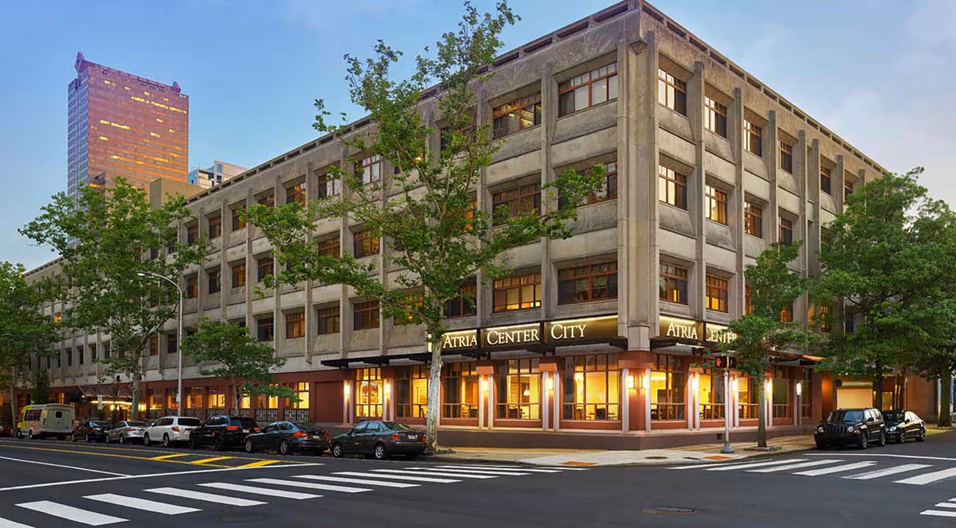 Exterior view of a multi-story building named Atria Center City at dusk, with trees lining the street and several parked cars along the curb.